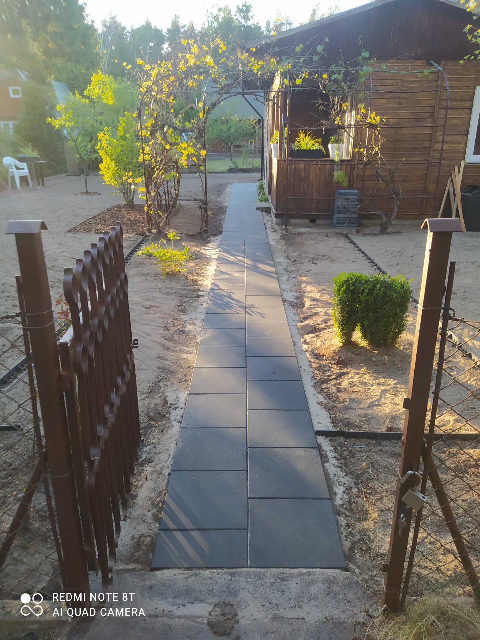 Ścieżka z szarych płyt prowadząca od metalowej bramki do drewnianego domku z werandą, nad którą rosną pnącza. W tle widoczne drzewa i budynek.
