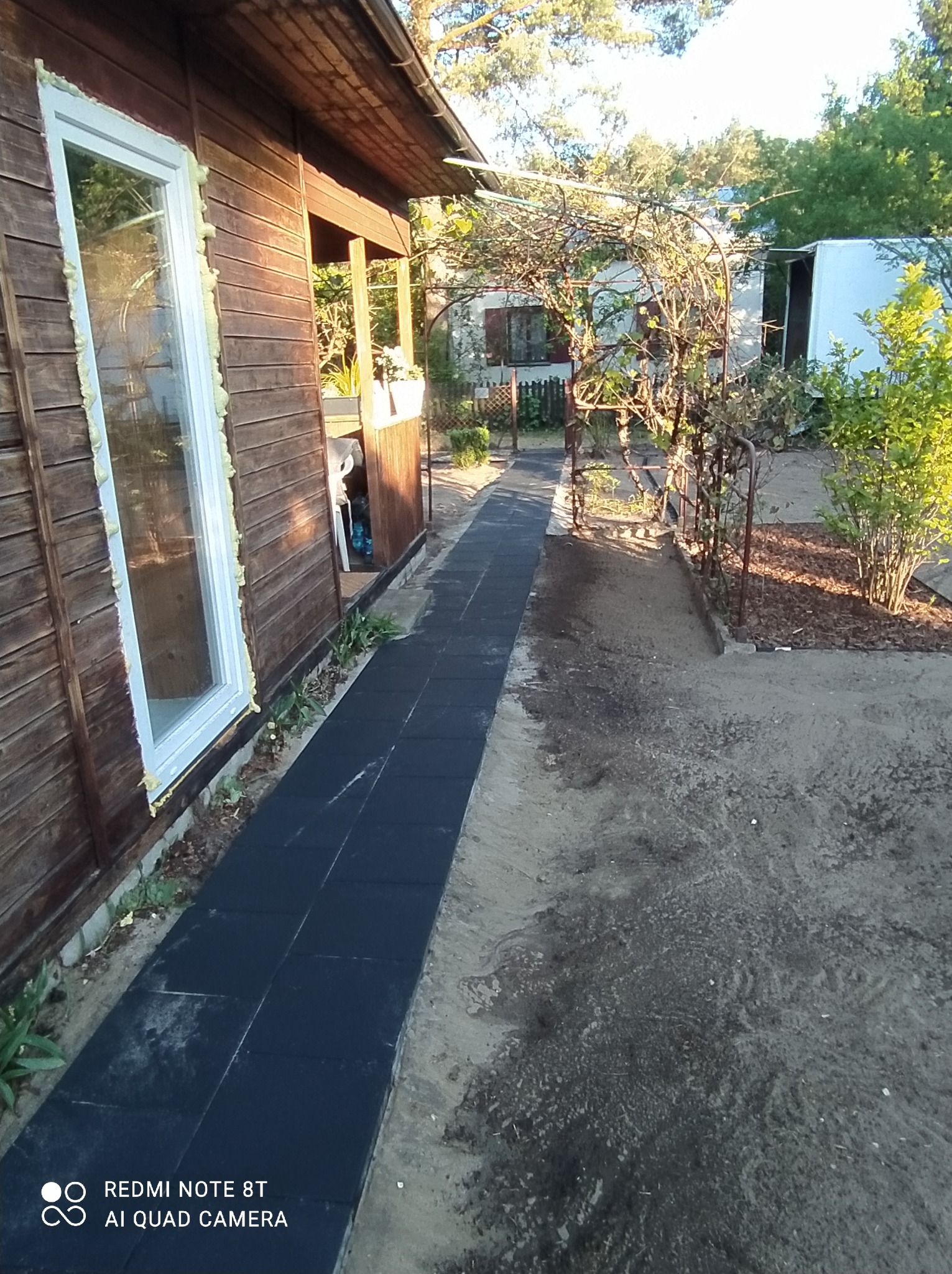 Ścieżka z czarnych, gumowych płyt prowadząca wzdłuż drewnianego domku w kierunku ogrodu z metalową pergolą i zielenią.