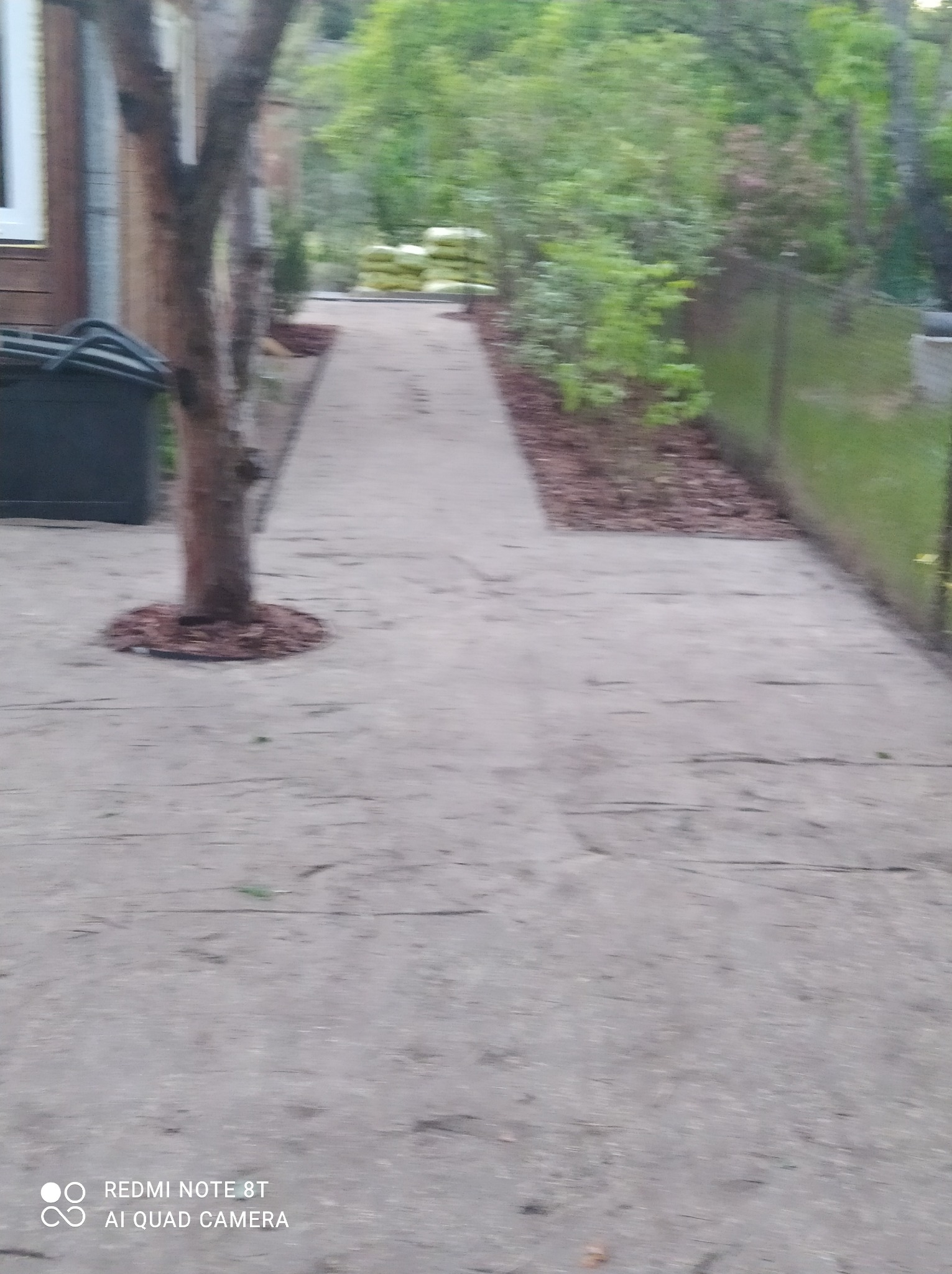 Ułożona szara nawierzchnia ścieżki ogrodowej z obrzeżami z kory, drzewo w centrum kadru, w tle worki z ziemią i zieleń.