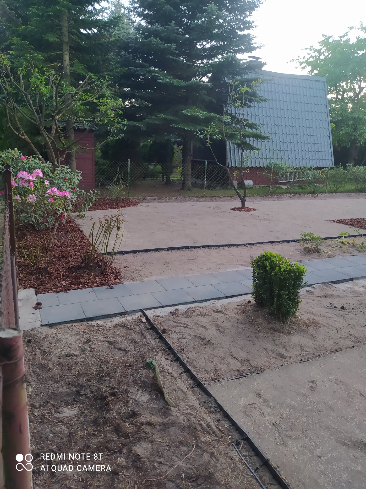 Przygotowany teren pod ścieżkę z kostki brukowej, widoczne obrzeża, podsypka i częściowo ułożona kostka, w tle dom z szarym dachem i drzewa.