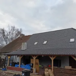 Dom z częściowo wymienionym pokryciem dachowym na ciemnoszarą dachówkę, widoczne okna dachowe i drewniana konstrukcja werandy, w tle drzewo bez liści i pochmurne niebo.