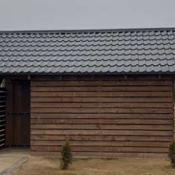 Mała, drewniana budowla z czarnym dachem z blachodachówki w Drezdenku, otoczona zielenią i małymi drzewkami iglastymi.