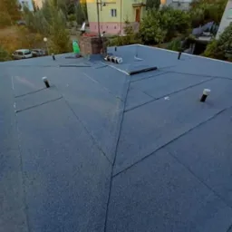 Nowe pokrycie dachowe z papy bitumicznej na dachu domu jednorodzinnego, widoczne komin i wentylacja, w tle zabudowa mieszkalna i zieleń.