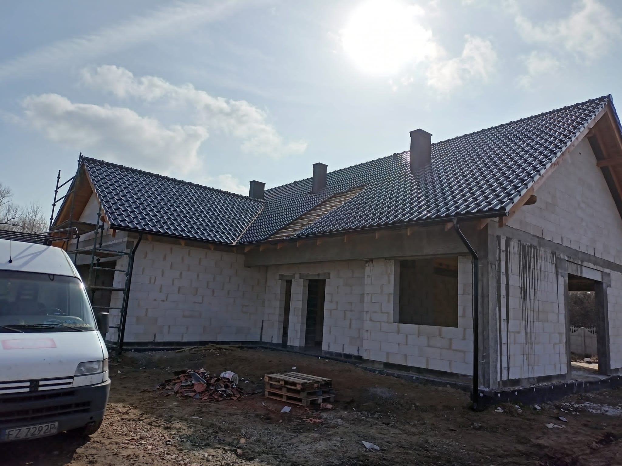 Niedokończony dom z częściowo położonym, ciemnoszarym dachem z dachówki ceramicznej, widoczne rusztowanie, zaparkowany biały bus i paleta z resztkami dachówek na ziemi.