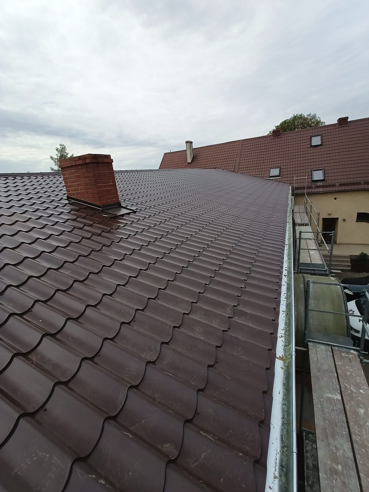 Nowy, brązowy dach z blachodachówki trapezowej, widoczny komin z cegły, rynna z zabrudzeniami i rusztowanie na tle pochmurnego nieba.