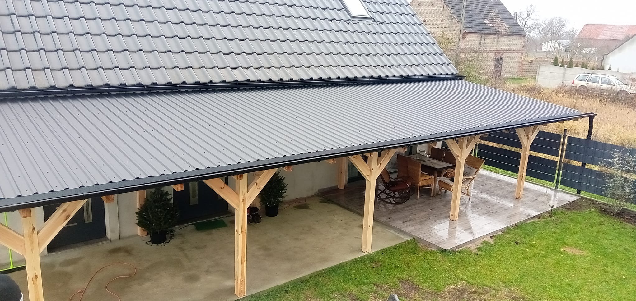 Widok z góry na zadaszony taras z drewnianą konstrukcją i metalowym dachem, przylegający do domu z szarym dachem ceramicznym. Widoczne rynny w kolorze czarnym. W tle budynki gospodarcze.