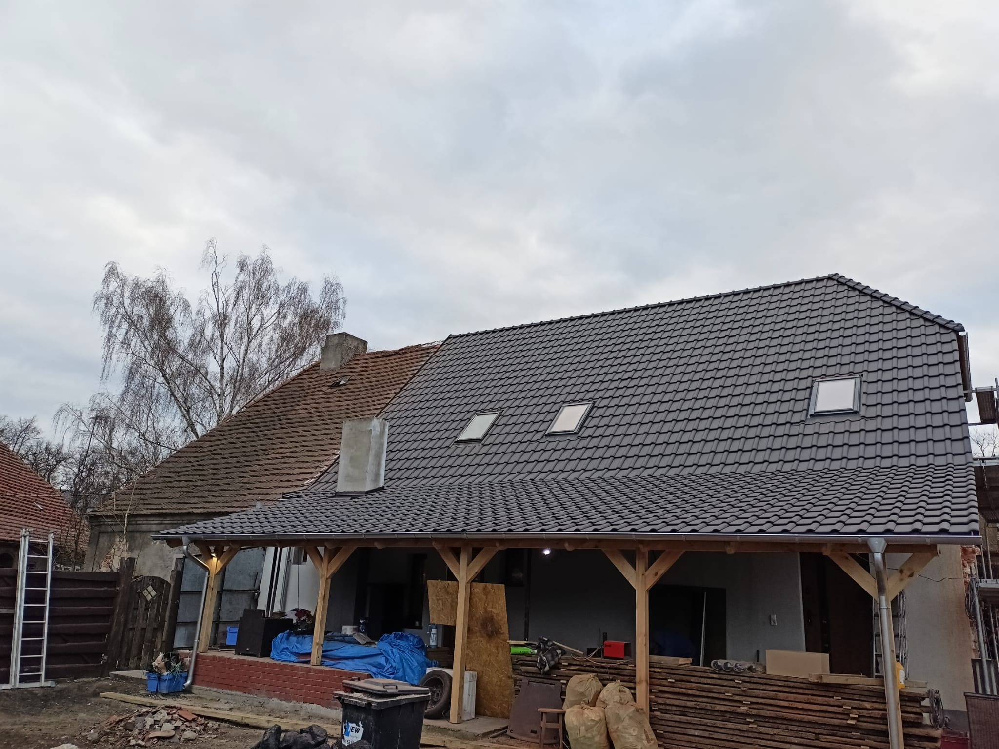 Dom z częściowo wymienionym pokryciem dachowym na ciemnoszarą dachówkę, widoczne okna dachowe i drewniana konstrukcja werandy, w tle drzewo bez liści i pochmurne niebo.