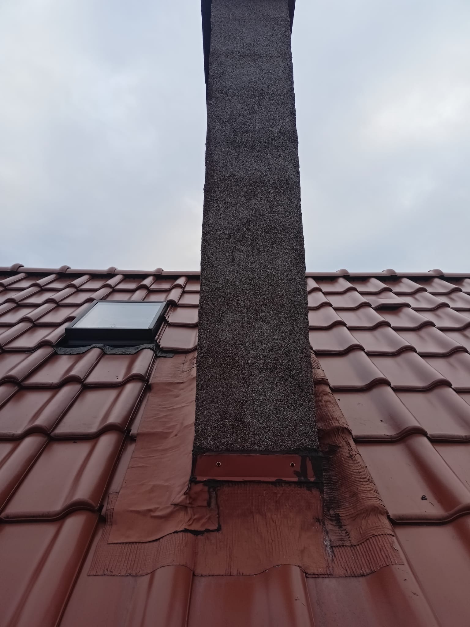 Czerwona dachówka ceramiczna z kominem i oknem dachowym; uszczelnienie komina wykonane z papy bitumicznej, widoczne ślady napraw.