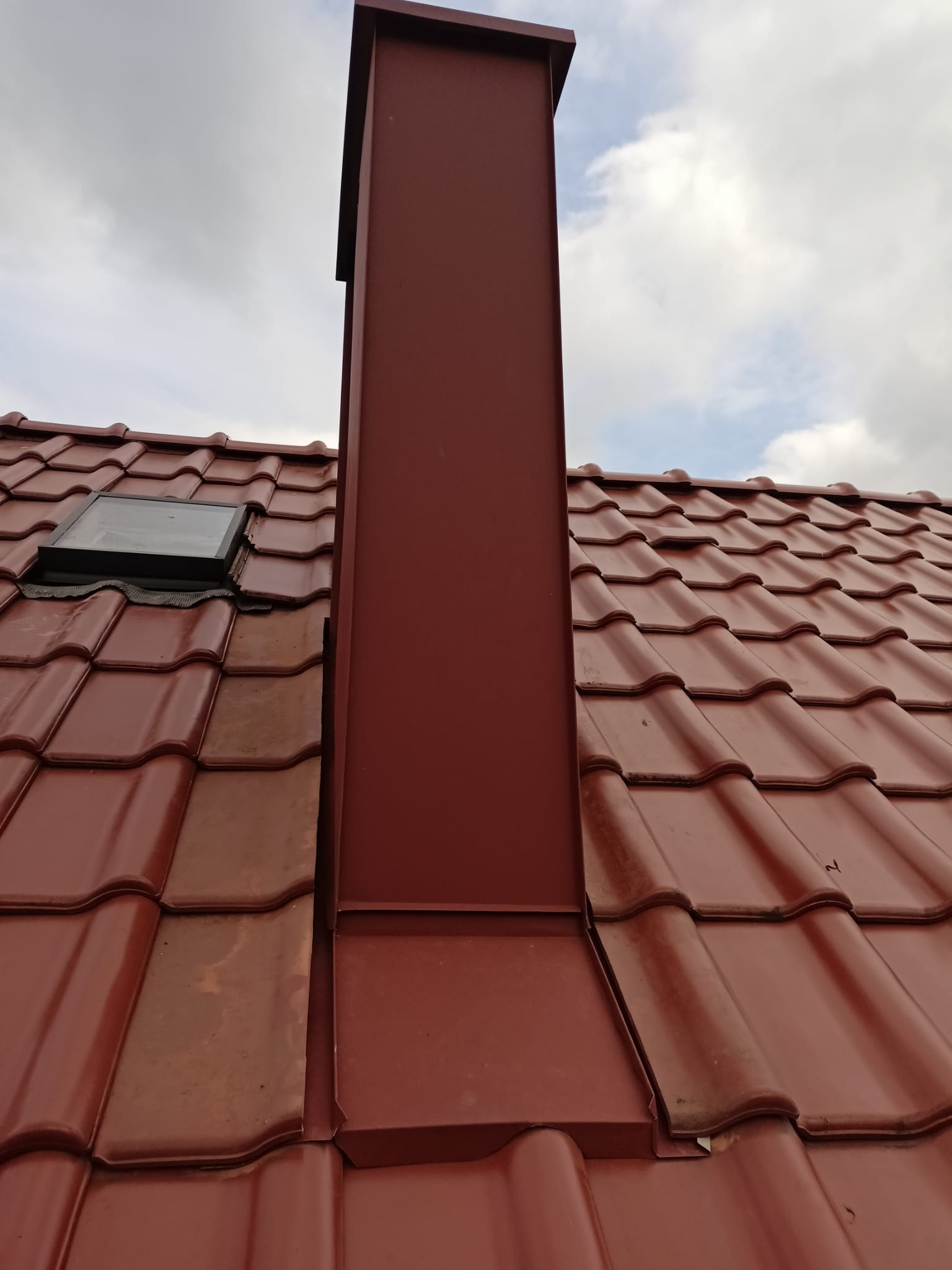 Czerwony dach z dachówki ceramicznej z nowym kominem w kolorze dopasowanym do dachu i oknem dachowym w lewym górnym rogu, na tle pochmurnego nieba.