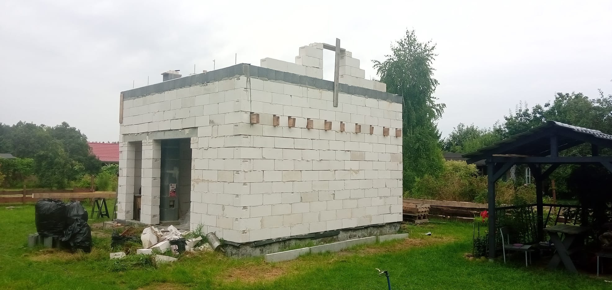 Budowa domu jednorodzinnego w stanie surowym otwartym z bloczków, widoczne drewniane elementy szalunku, otoczenie zieleni.