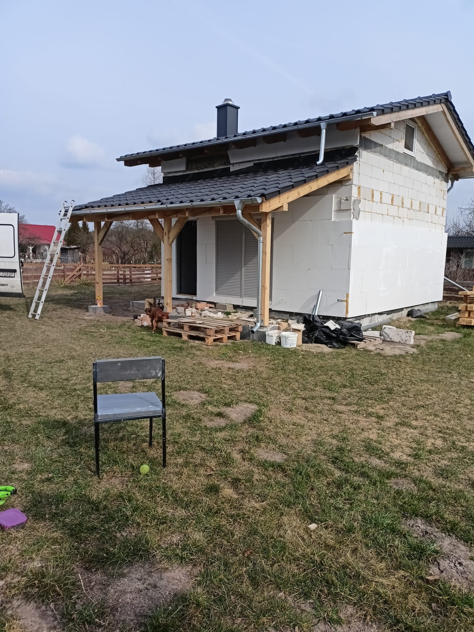 Dom w trakcie budowy z widocznym dachem z czarnej dachówki, zamontowanymi rynnami, częściowo ocieplonymi ścianami, drewnianą konstrukcją werandy i drabiną opartą o dach, na pierwszym planie krzesło...
