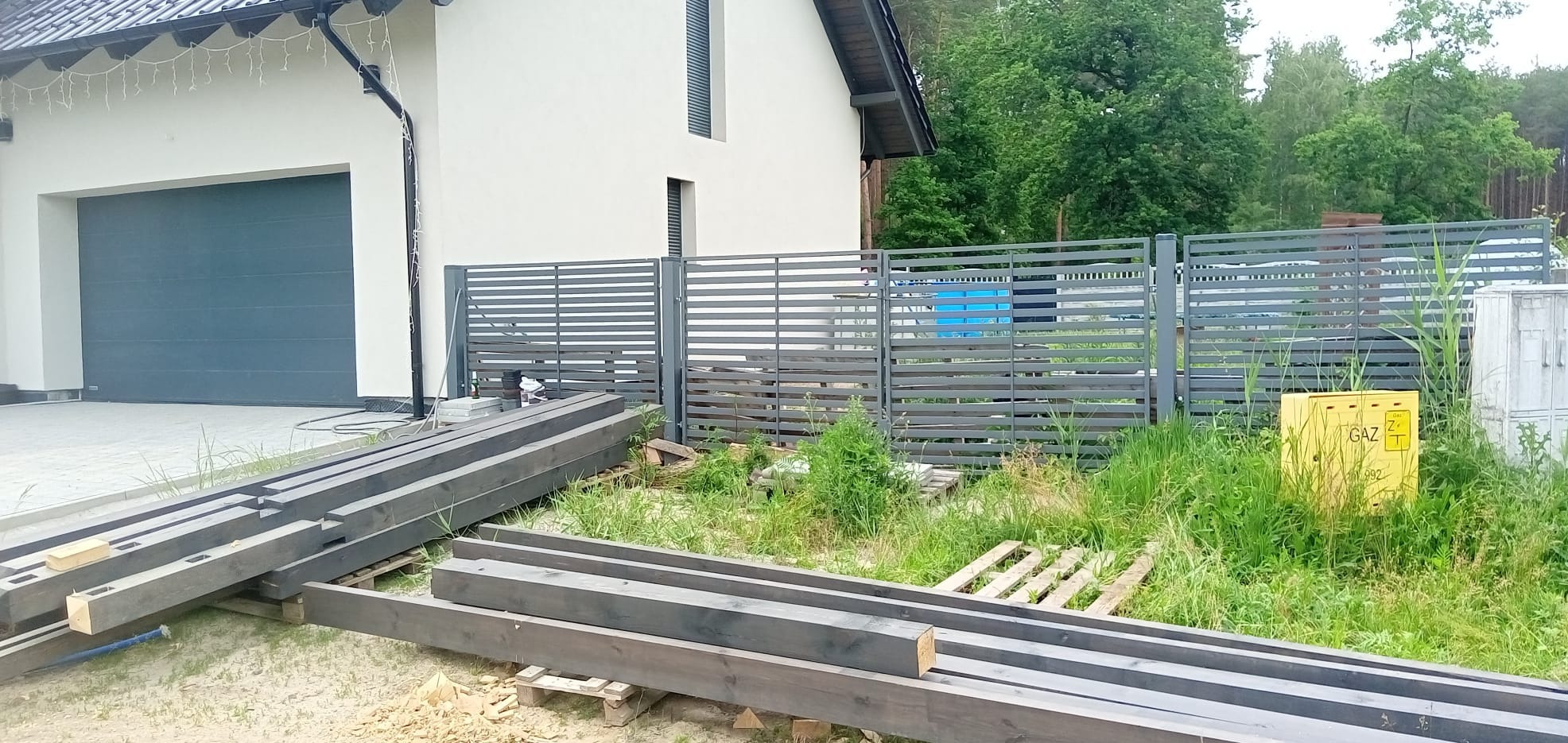 Przygotowane, impregnowane drewniane belki leżące na paletach przed nowoczesnym domem z szarą, poziomą metalową bramą i skrzynką gazową w trawie.