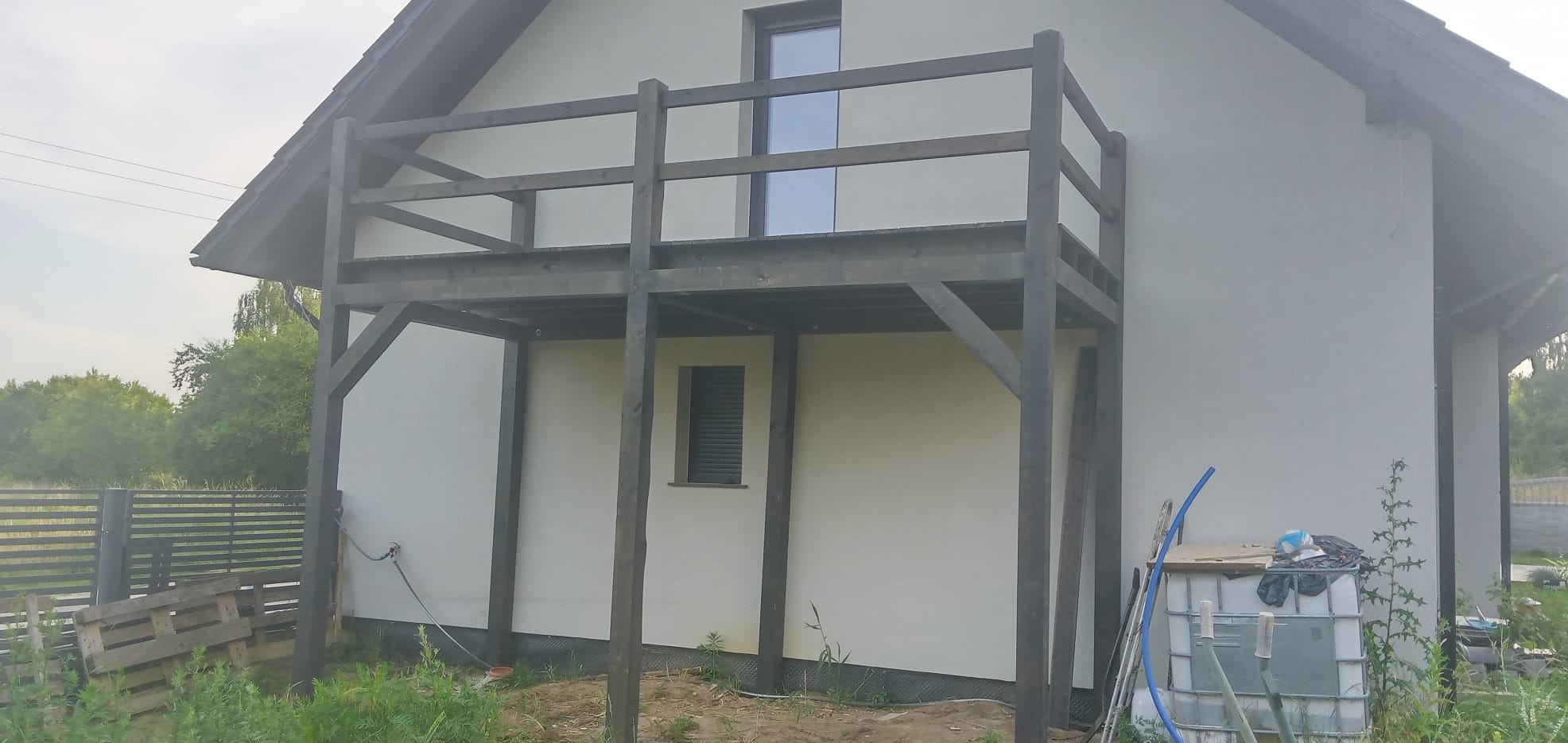 Drewniana konstrukcja balkonu przylegająca do ściany domu z widocznymi elementami dachu, rynną, oknami oraz zbiornikiem na wodę deszczową i paletami w tle.