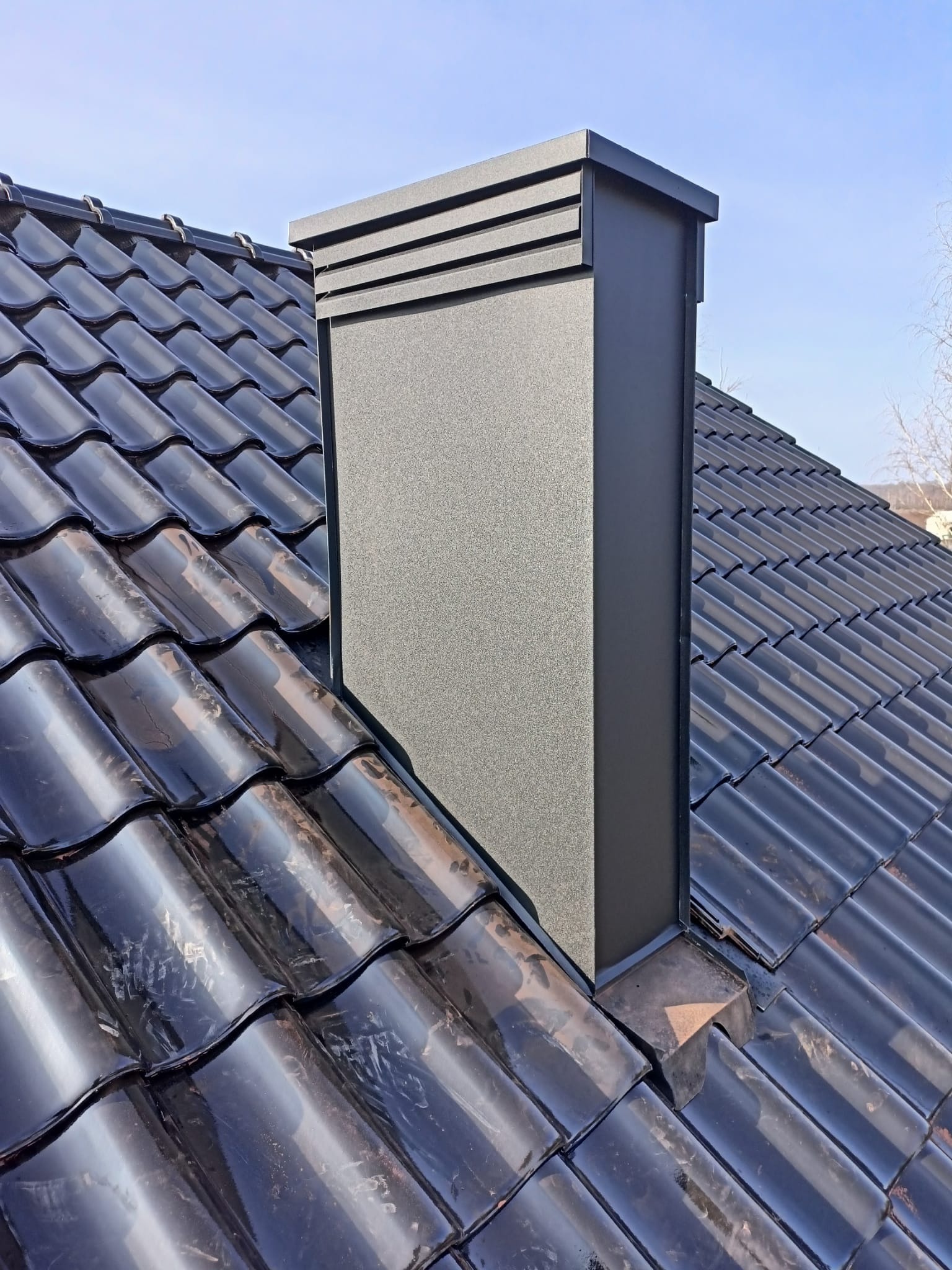 Czarny, nowoczesny komin wentylacyjny na dachu z ciemnej, błyszczącej dachówki ceramicznej, widok z dołu pod kątem, na tle błękitnego nieba.