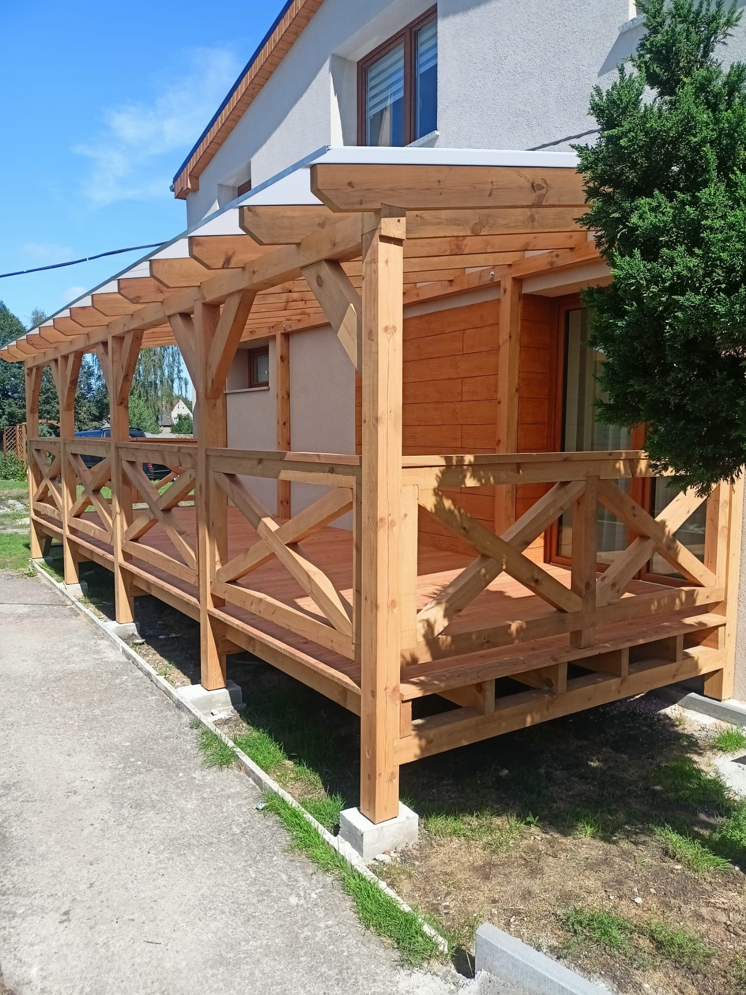 Drewniana pergola z balustradą w kształcie krzyży, przylegająca do budynku z jasną elewacją, widok z zewnątrz w słoneczny dzień.