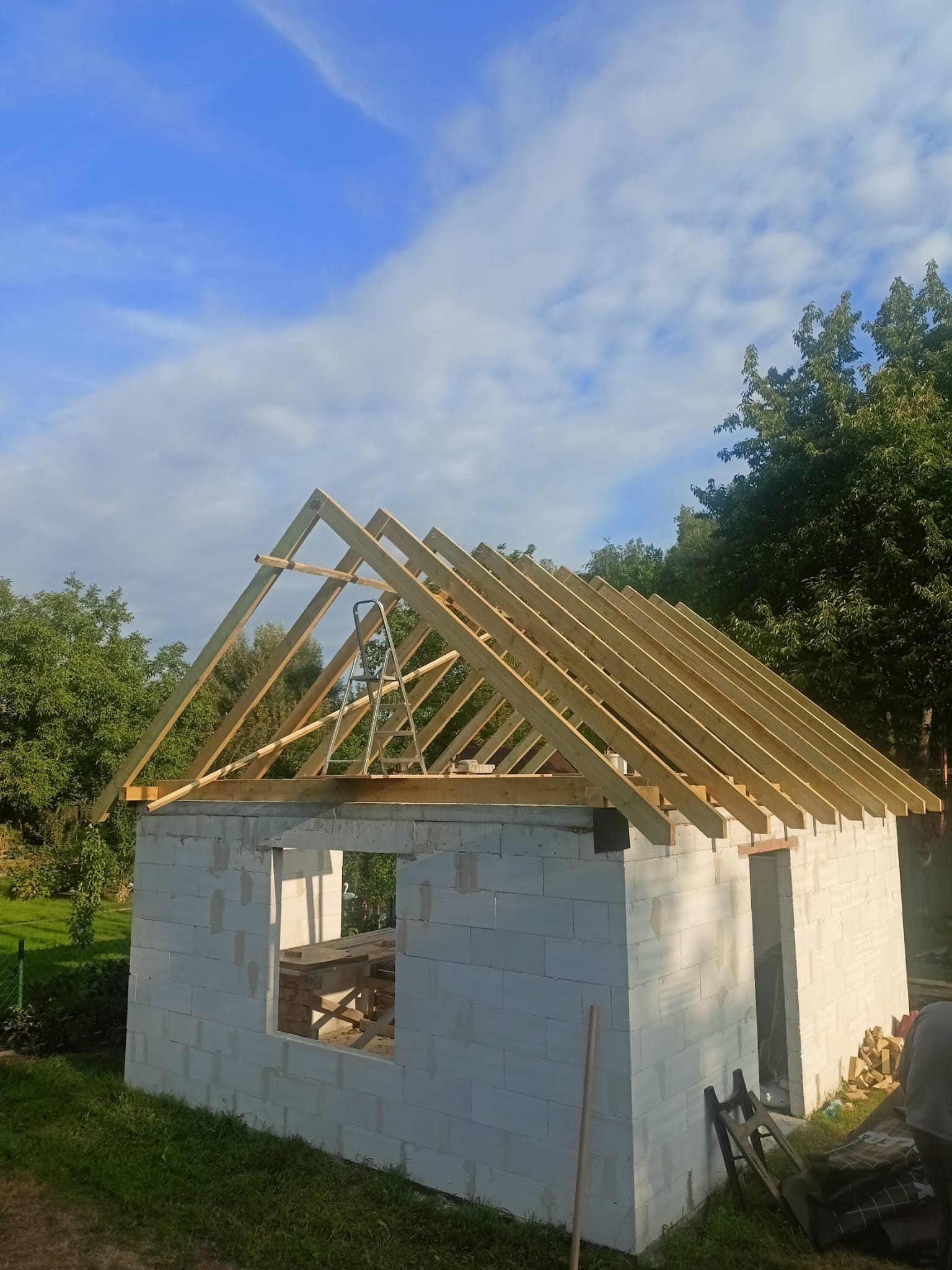 Konstrukcja drewniana dachu w trakcie budowy na budynku z białych bloczków, z drabiną opartą o krokwie na tle błękitnego nieba i zieleni.