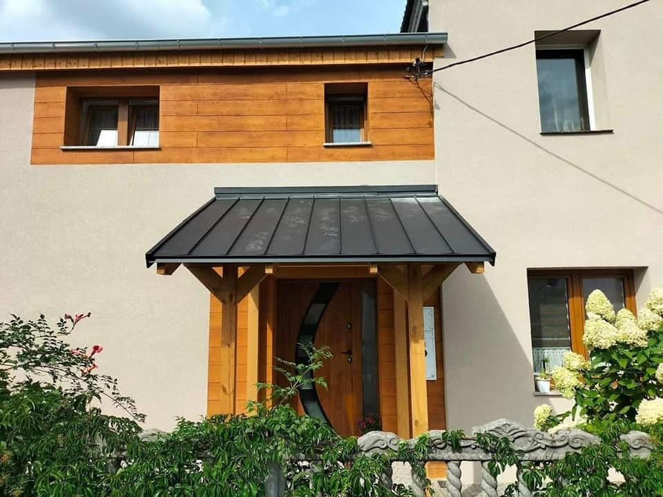 Dwukondygnacyjny dom z drewnianą elewacją na górze i beżową na dole, z drewnianym zadaszeniem nad wejściem pokrytym blachą w kolorze grafitowym.
