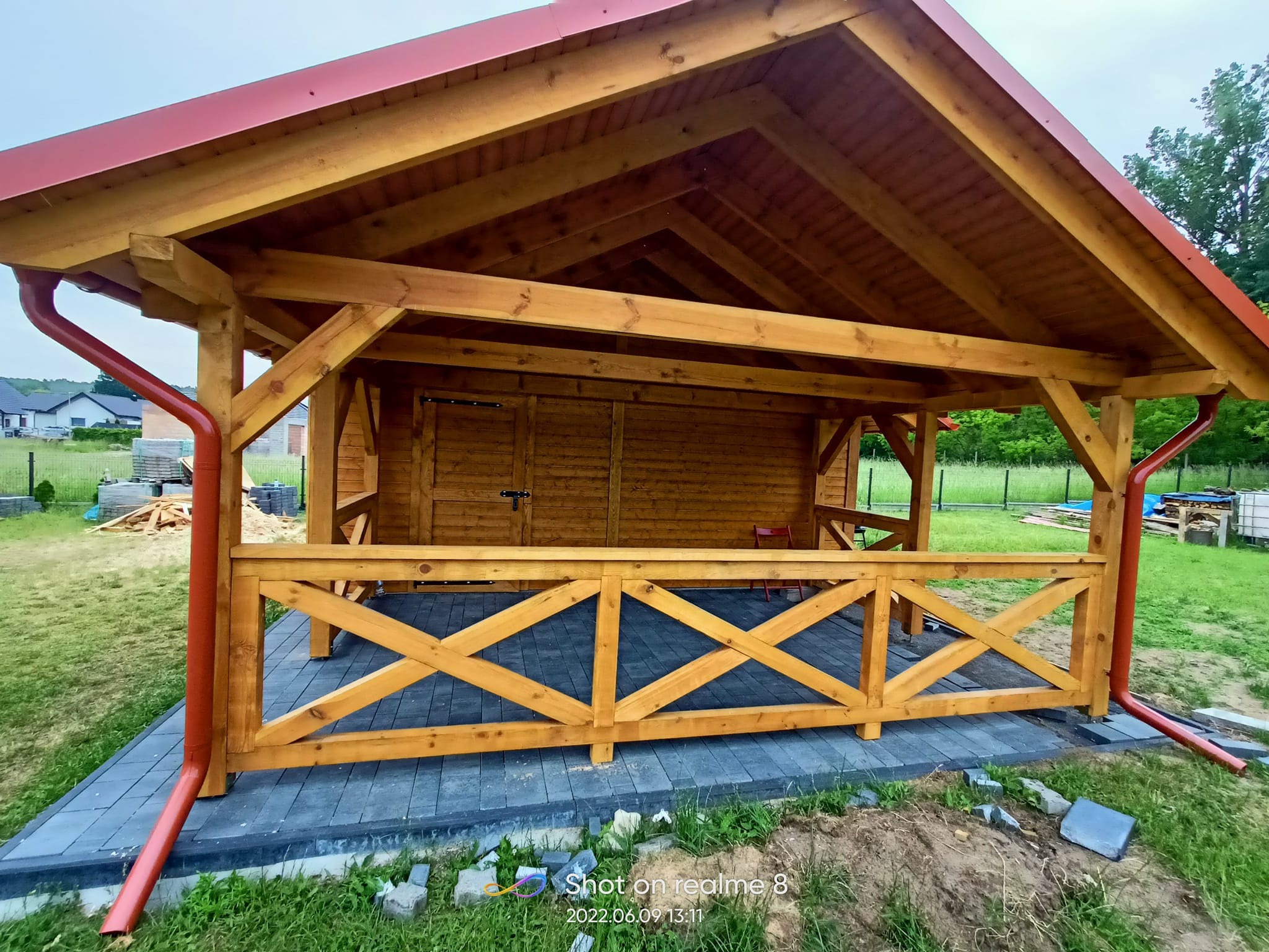 Drewniana altana z czerwonymi rynnami i dachem, widok z zewnątrz na drewniane balustrady i wyłożony kostką taras.