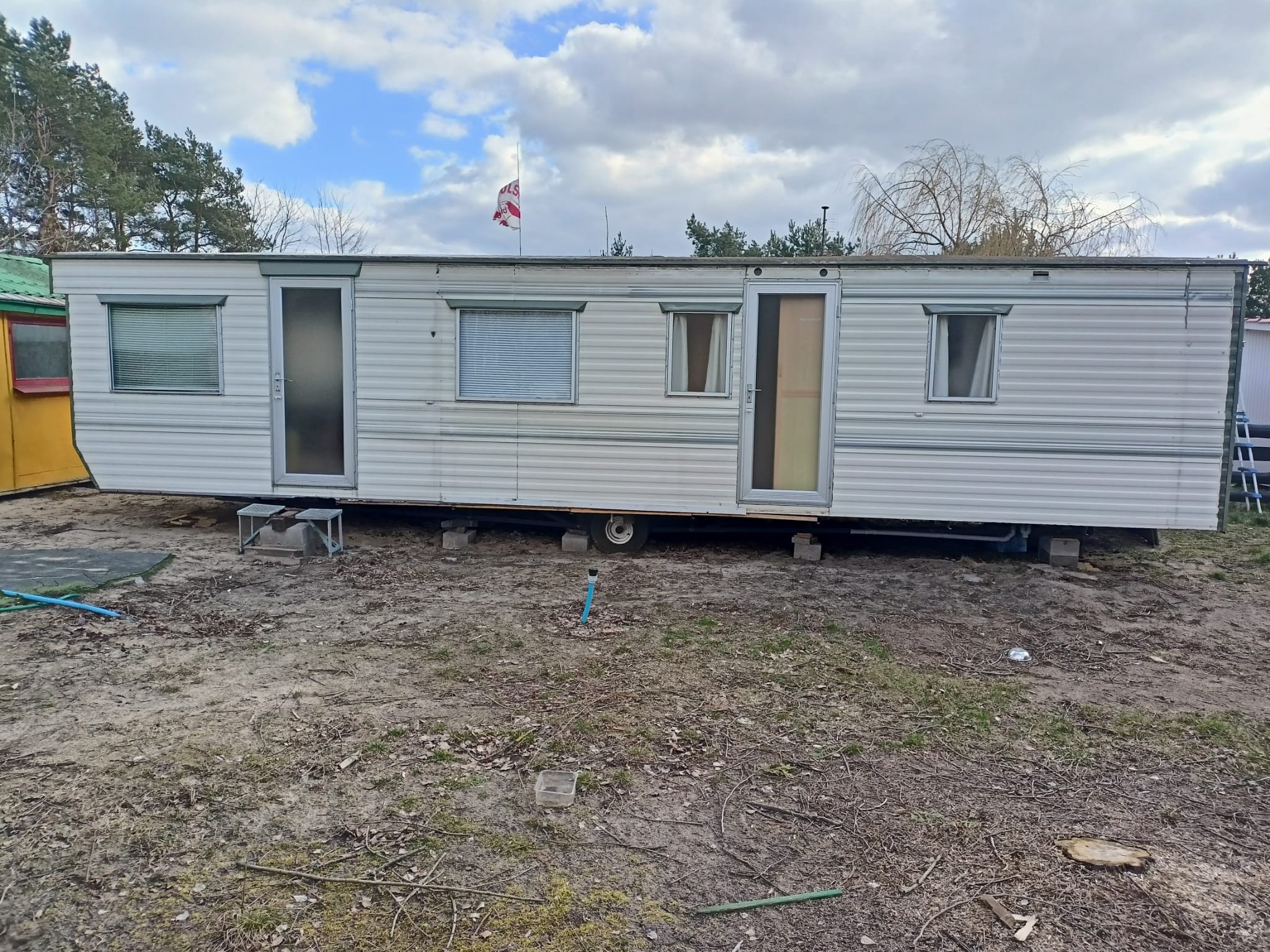 Długi, parterowy, biały dom mobilny z szarymi akcentami, ustawiony na betonowych blokach, z flagą na dachu, na tle pochmurnego nieba i drzew