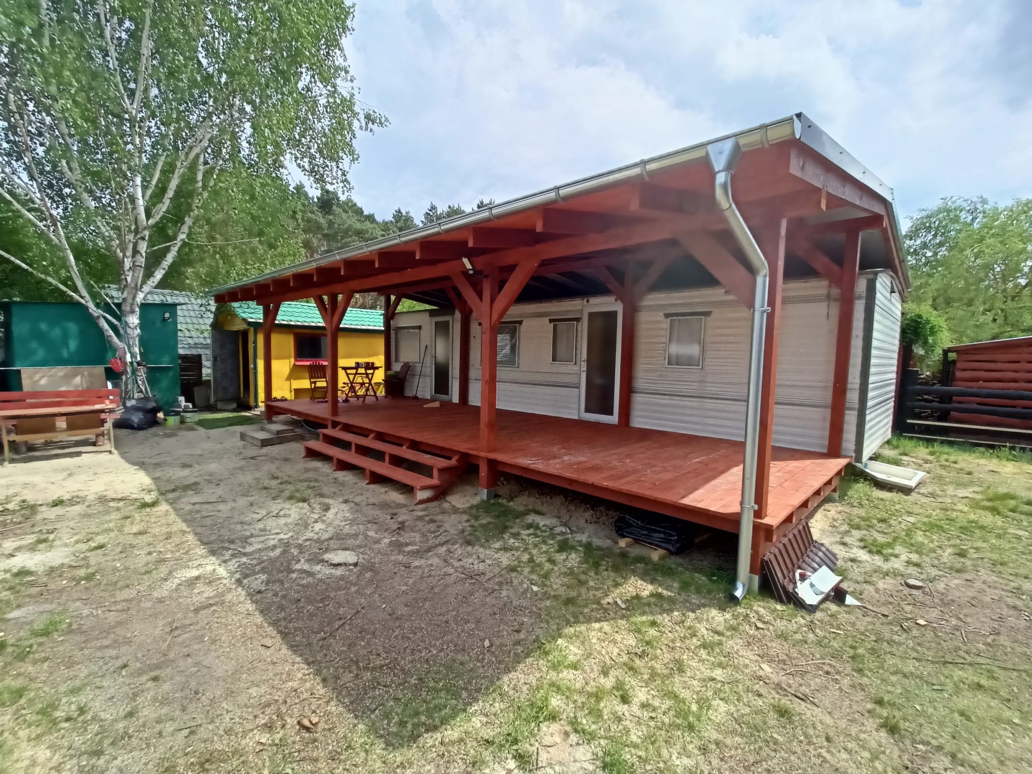 Szary domek mobilny z dobudowanym, zadaszonym tarasem z pomalowanym na czerwono drewnianym szkieletem, widoczne rynny spustowe.