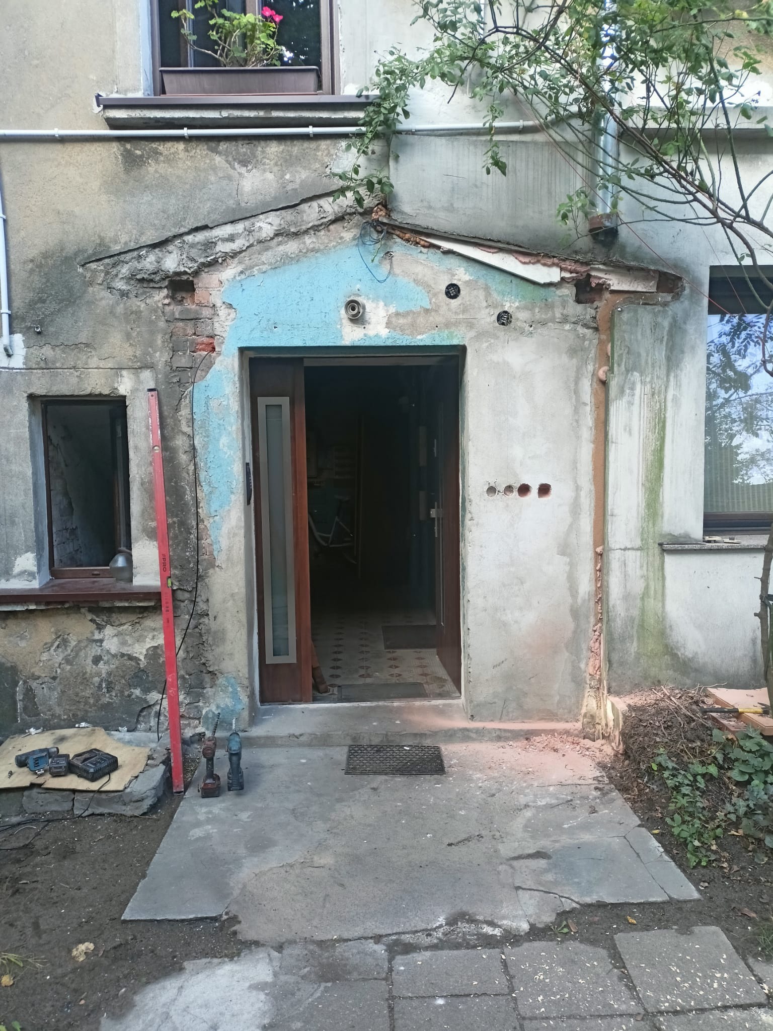 Wejście do budynku w trakcie remontu, z odsłoniętymi fragmentami muru i niebieskim malowaniem pod okapem, narzędzia i poziomica leżące obok.