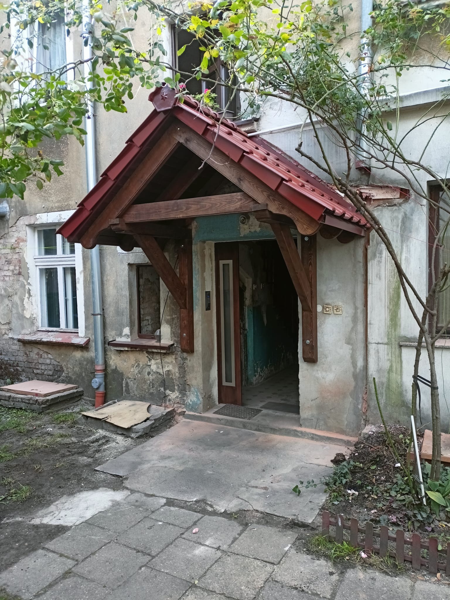 Wejście do budynku z drewnianym zadaszeniem krytym czerwoną dachówką, widoczne ślady zniszczeń na elewacji i chodniku, fragment rynny spustowej po lewej stronie.