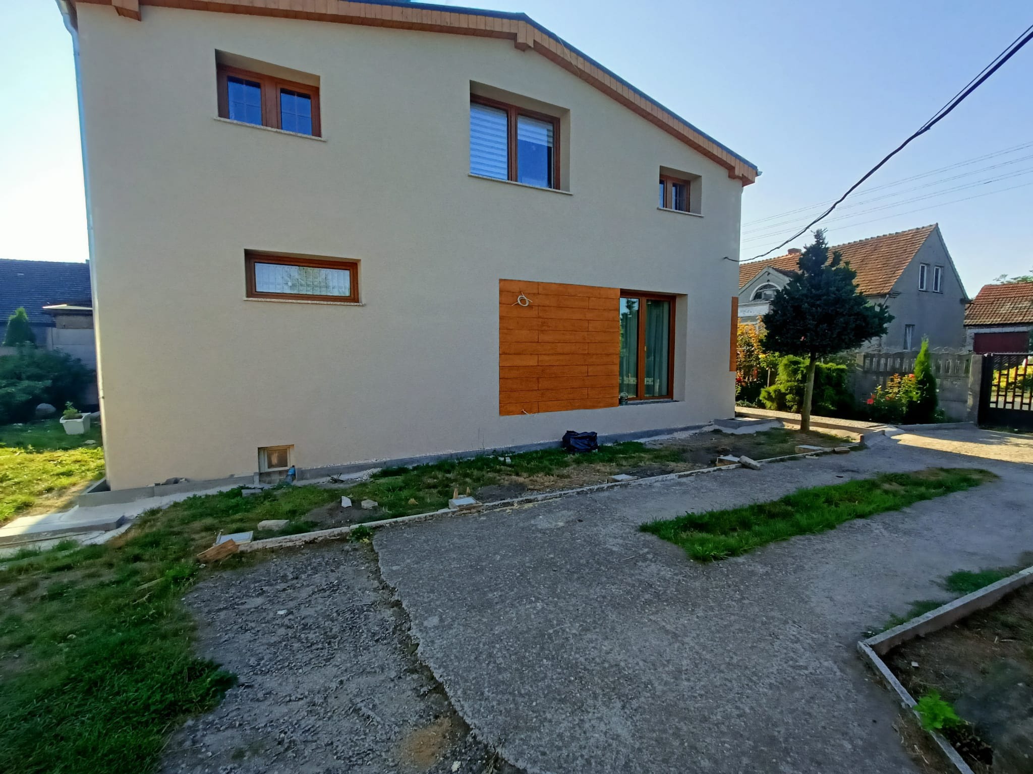 Nowoczesny dom z beżową elewacją, drewnianymi ramami okiennymi i dekoracyjnym panelem z drewna na ścianie, widok z zewnątrz na podwórze z betonowym podjazdem i trawą.