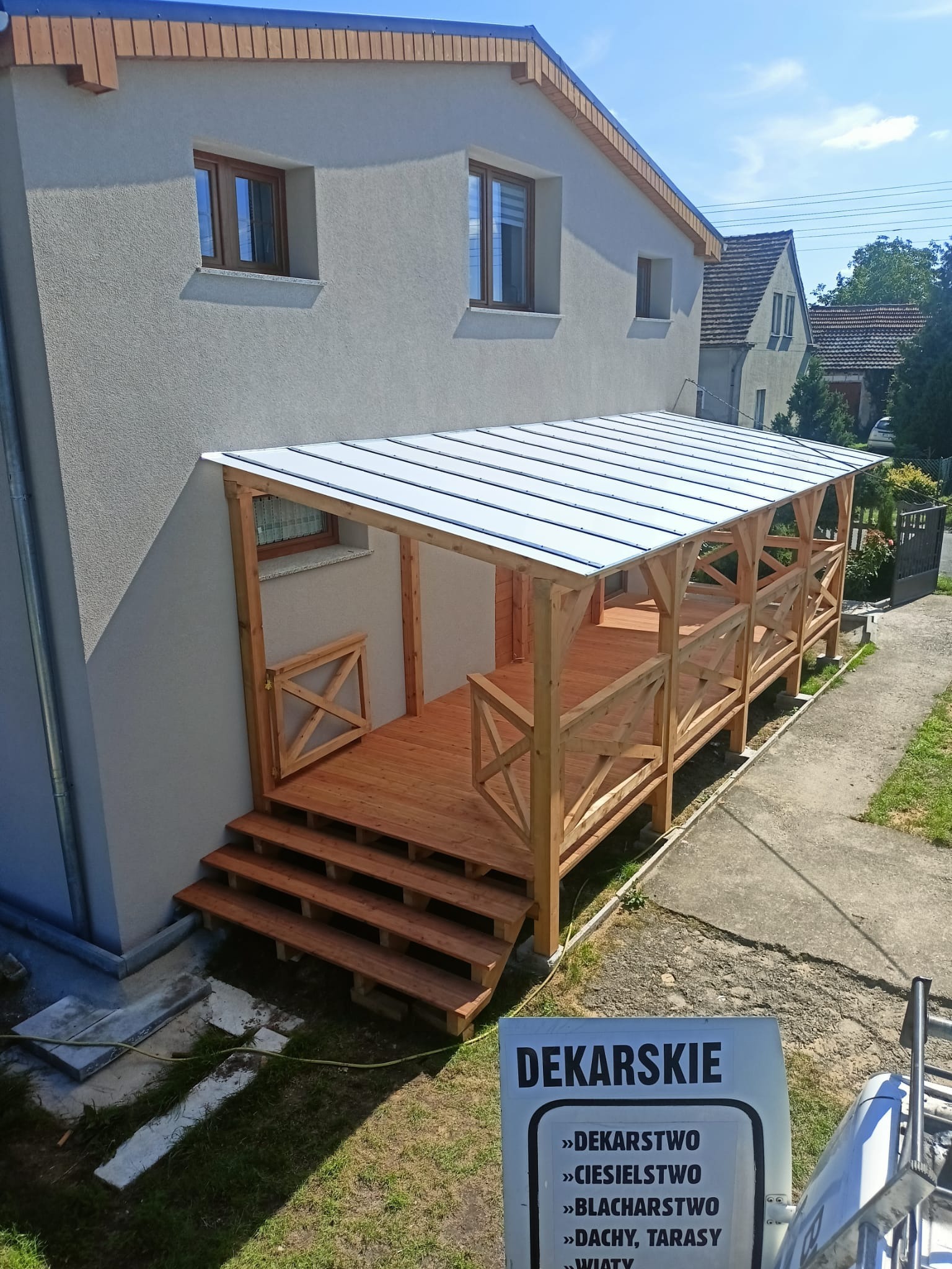 Drewniana weranda z zadaszeniem z blachy trapezowej, dobudowana do domu, widok z góry. Na pierwszym planie reklama usług dekarskich.
