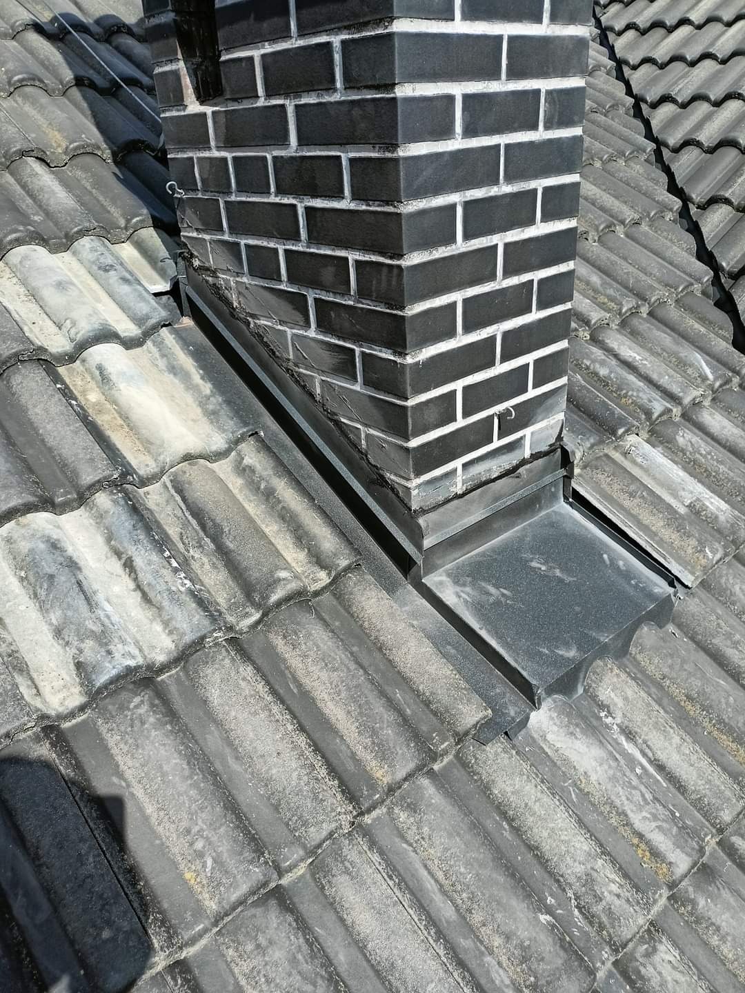 Czarny komin z cegły klinkierowej wystający z dachu pokrytego szarą dachówką ceramiczną. Obróbka blacharska wokół komina.