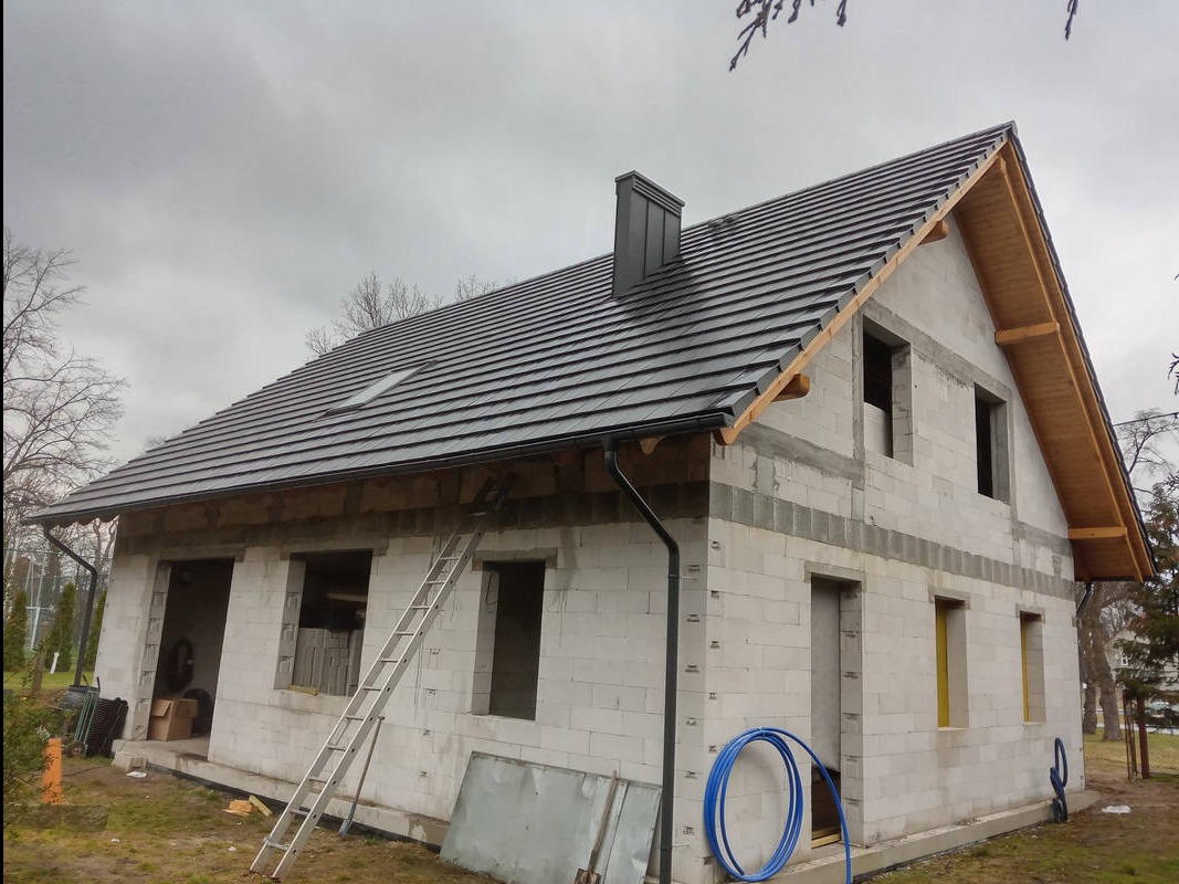 Dom w trakcie budowy z nowym, ciemnym dachem z dachówką, widoczne rynny i komin, drabina oparta o ścianę, stan surowy otwarty