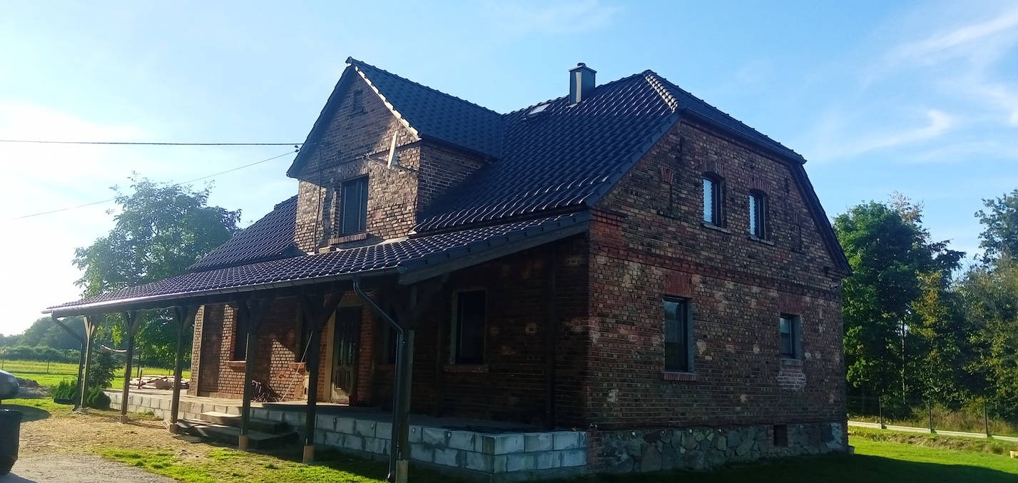 Dom z ciemnej cegły z nowym, ciemnym dachem i rynnami, widok z zewnątrz w słoneczny dzień.