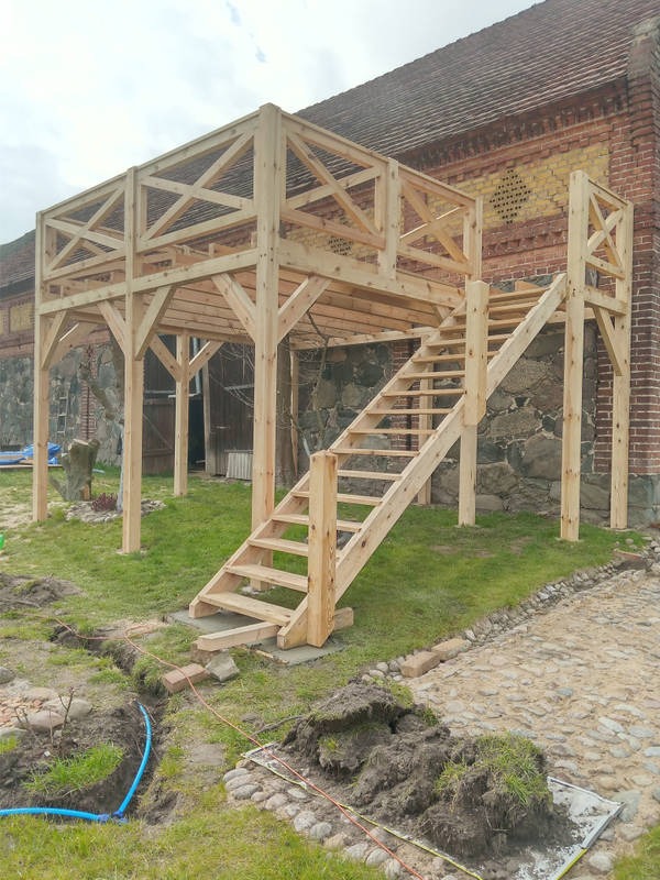 Drewniana konstrukcja tarasu z schodami, w trakcie budowy, z widocznym fundamentem i częściowo wykopaną ziemią, na tle ceglanego budynku.
