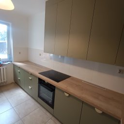 Kamil Żeliszczak - Nowoczesna kuchnia z oliwkowymi szafkami z półokrągłymi uchwytami, drewnianym blatem, czarną płytą indukcyjną i piekarnikiem w zabudowie, widok na grzejnik pod oknem.