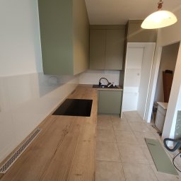 Kamil Żeliszczak - Wnętrze kuchni w trakcie montażu: drewniany blat z wbudowaną płytą grzewczą, zielone szafki wiszące, widoczny odkurzacz i elementy wykończeniowe na podłodze.