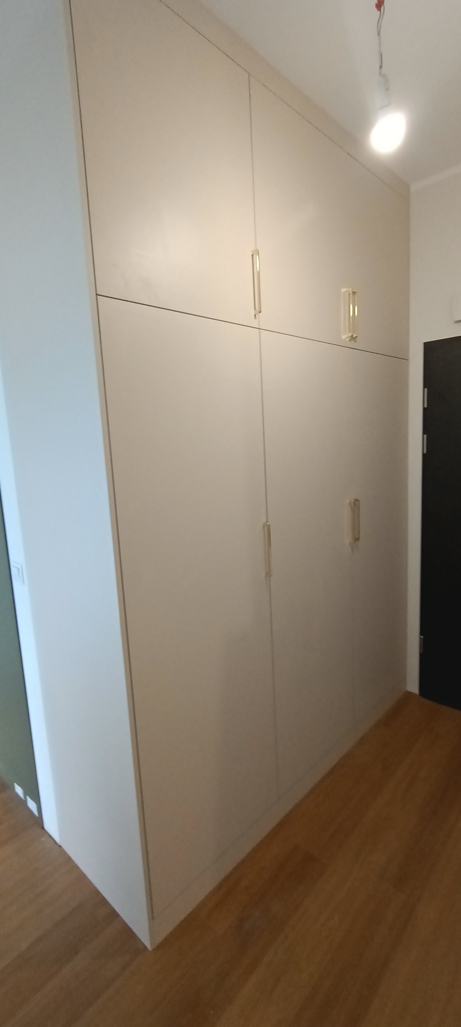 Wysoka, zabudowana szafa wnękowa z jasnymi, gładkimi frontami i minimalistycznymi, pionowymi uchwytami, dopasowana do skosu ściany w przedpokoju.