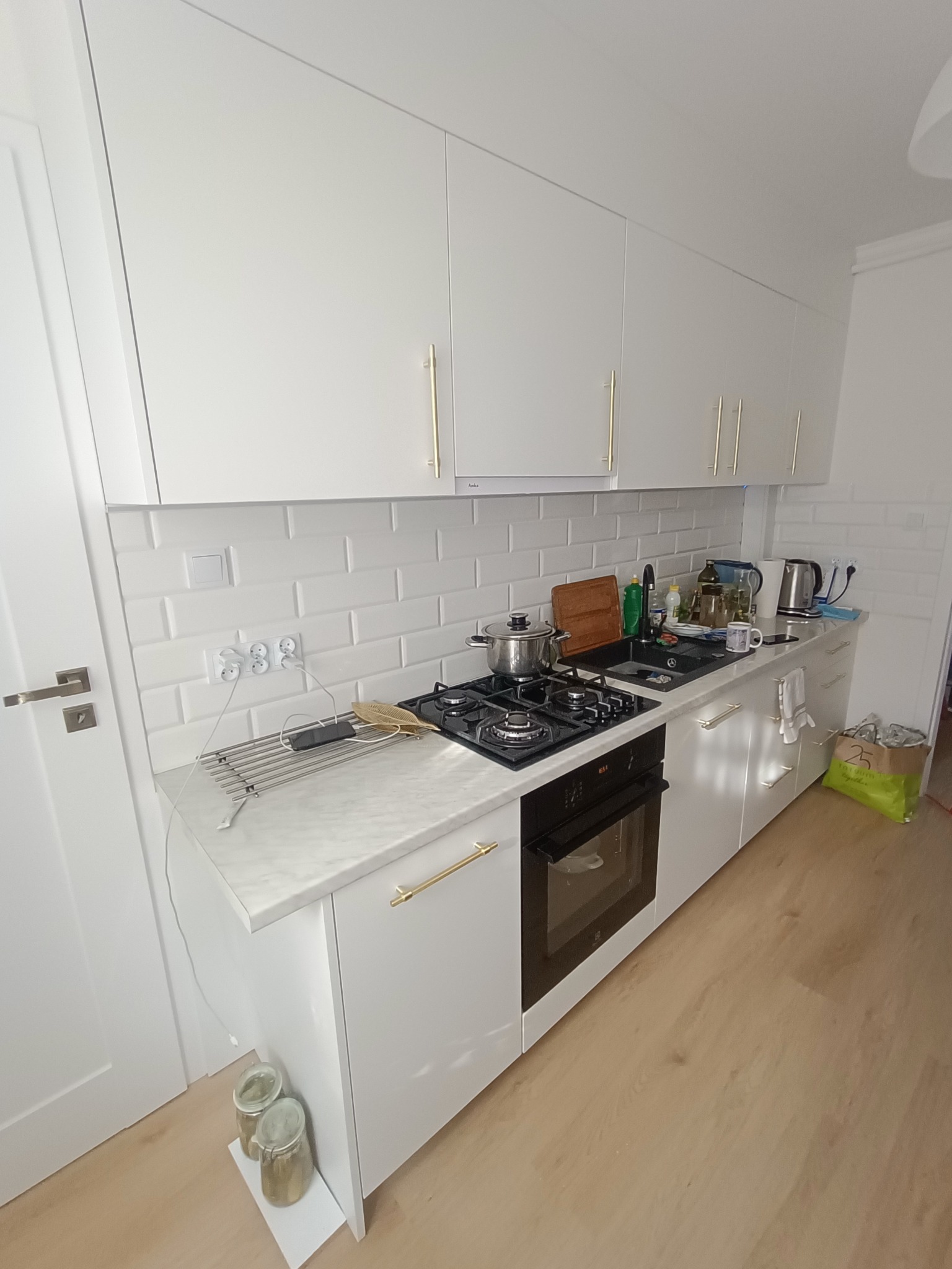 Biała kuchnia z białymi szafkami, złotymi uchwytami, czarnym blatem, płytą gazową i piekarnikiem; widoczne akcesoria kuchenne i białe płytki na ścianie.