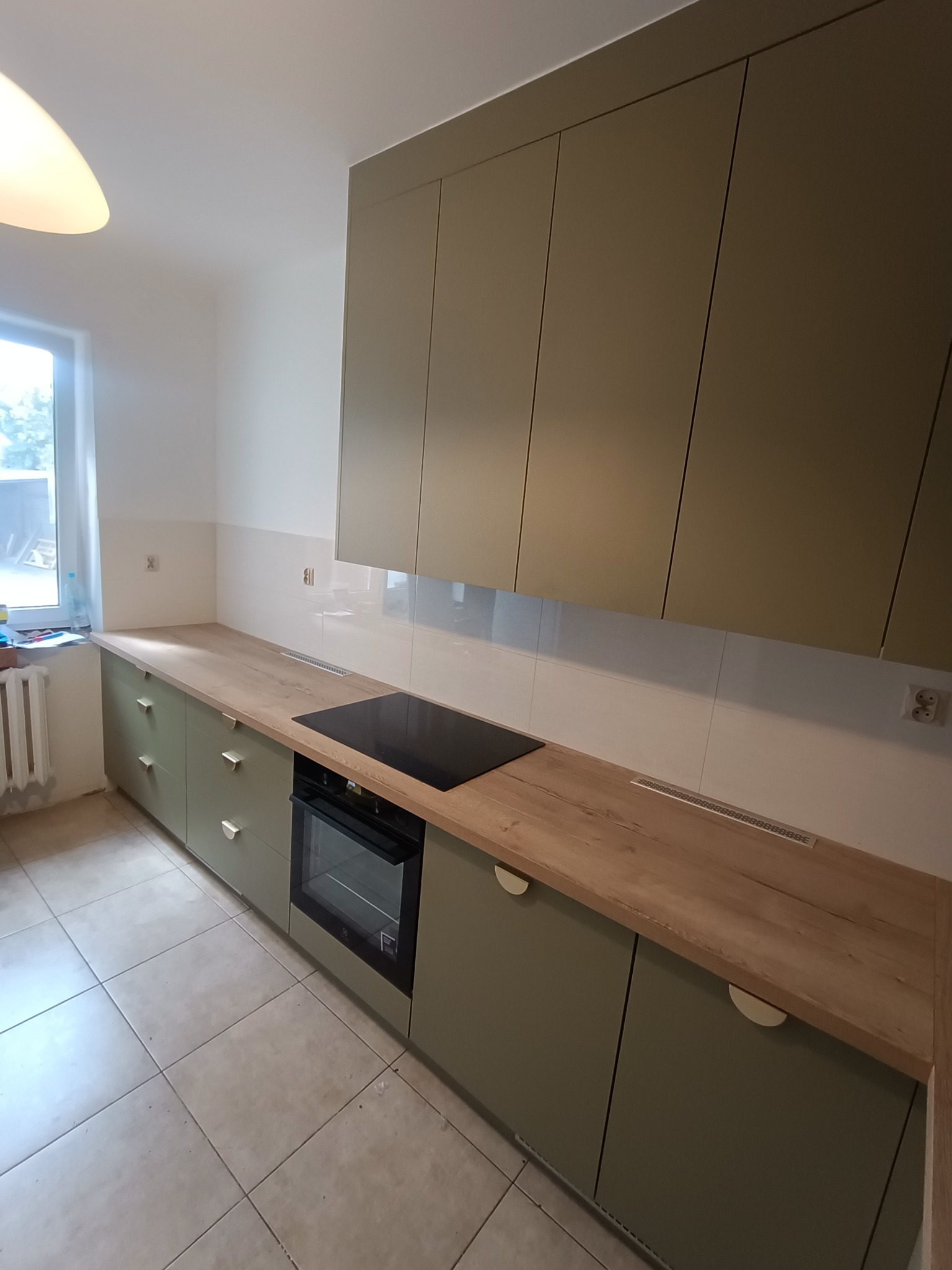 Nowoczesna kuchnia z oliwkowymi szafkami z półokrągłymi uchwytami, drewnianym blatem, czarną płytą indukcyjną i piekarnikiem w zabudowie, widok na grzejnik pod oknem.