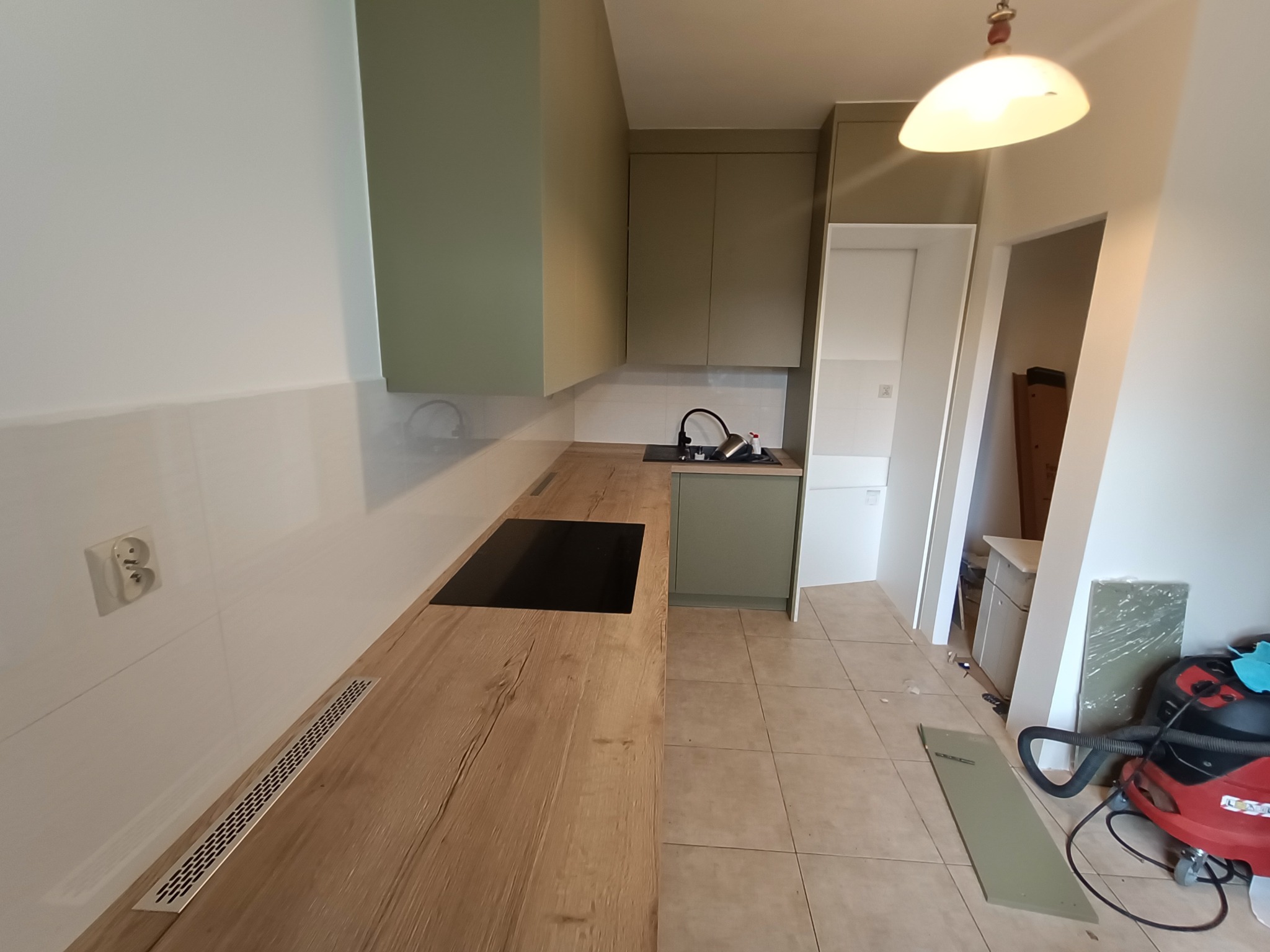 Wnętrze kuchni w trakcie montażu: drewniany blat z wbudowaną płytą grzewczą, zielone szafki wiszące, widoczny odkurzacz i elementy wykończeniowe na podłodze.