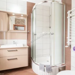 Jasna łazienka z kabiną prysznicową z zaokrąglonym szkłem, umywalką z szafką w beżowym odcieniu, grzejnikiem drabinkowym i brązowymi akcentami na ścianach.