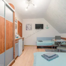 Pokój w pensjonacie z dwoma łóżkami z turkusową pościelą, stolikiem z szarymi krzesłami, telewizorem na ścianie i dekoracyjną kotwicą, oraz aneksem kuchennym z lodówką.