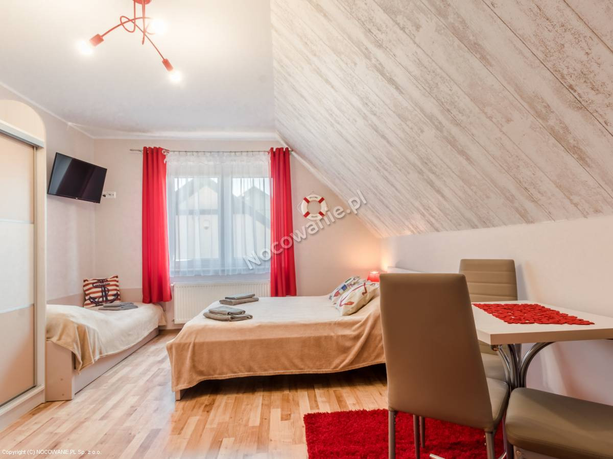 Pokój w stylu marynistycznym z dwoma łóżkami, stołem i krzesłami, dekoracjami w postaci koła ratunkowego i poduszki z kotwicą.