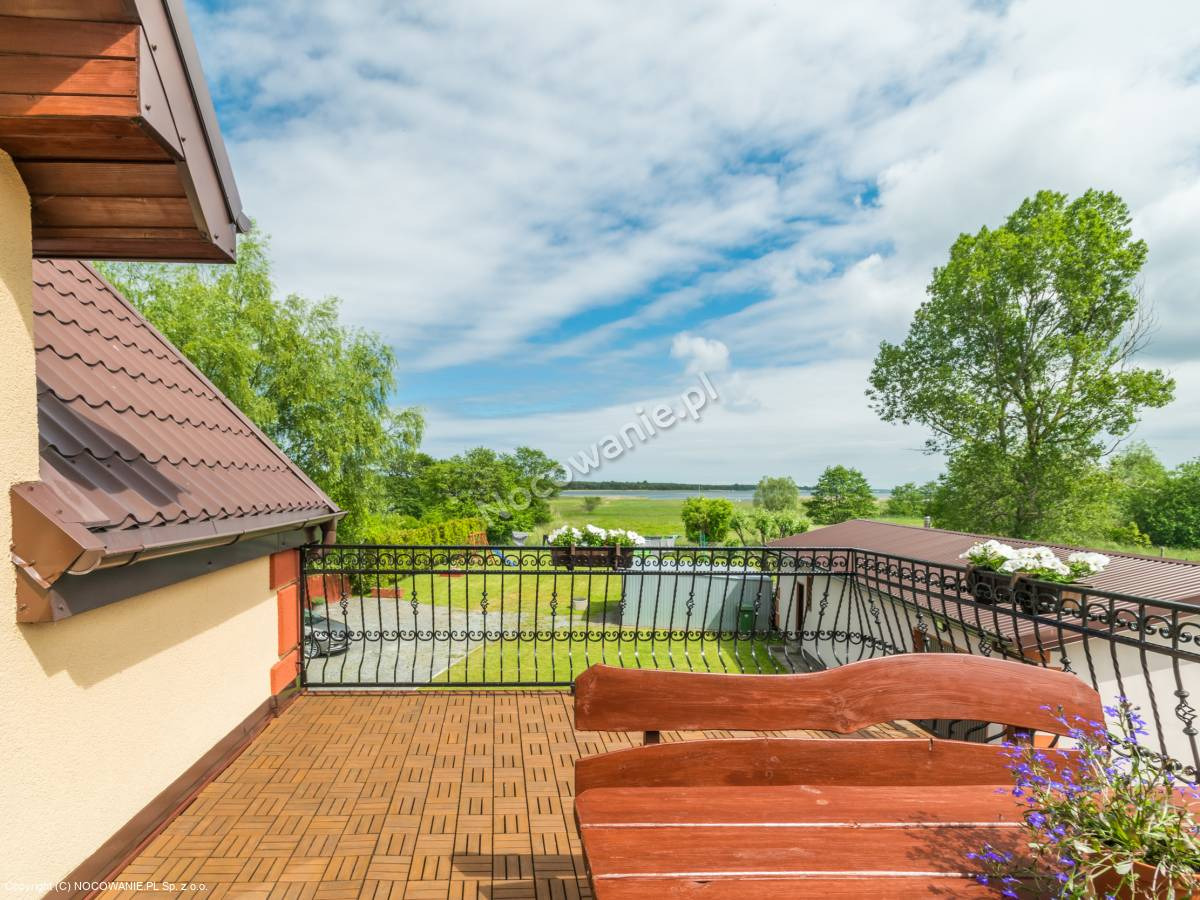 Widok z balkonu na jezioro, drewniany stół z kwiatami na pierwszym planie, ozdobna metalowa balustrada.