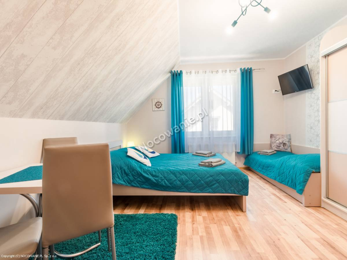 Pokój w pensjonacie z dwoma łóżkami przykrytymi turkusowymi narzutami, turkusowy dywanik, drewniana podłoga i skośny sufit z boazerią.