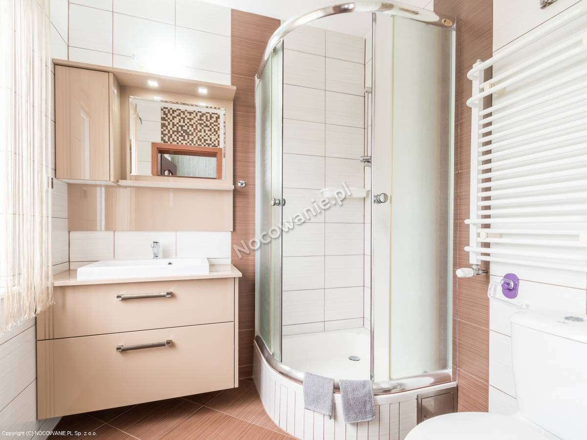 Jasna łazienka z kabiną prysznicową z zaokrąglonym szkłem, umywalką z szafką w beżowym odcieniu, grzejnikiem drabinkowym i brązowymi akcentami na ścianach.