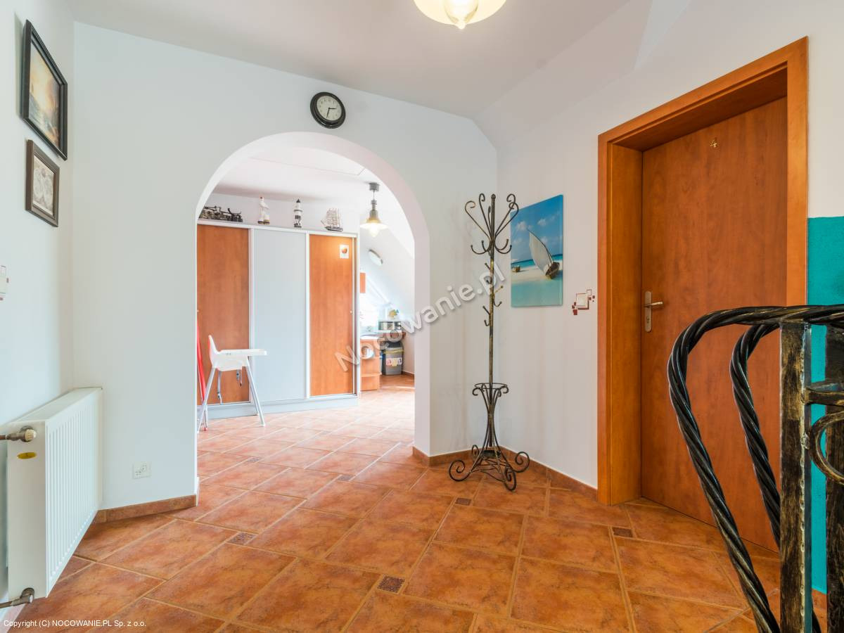 Hall w pensjonacie z terakotową podłogą, w tle widoczne szafy przesuwne i krzesełko dla dziecka, na ścianie wieszak na ubrania i obraz z motywem żaglówki.