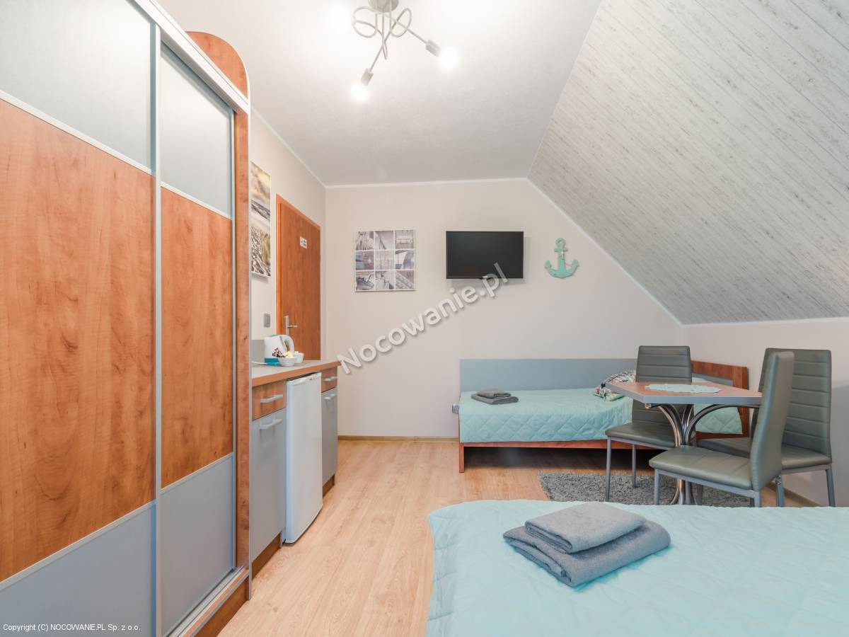 Pokój w pensjonacie z dwoma łóżkami z turkusową pościelą, stolikiem z szarymi krzesłami, telewizorem na ścianie i dekoracyjną kotwicą, oraz aneksem kuchennym z lodówką.