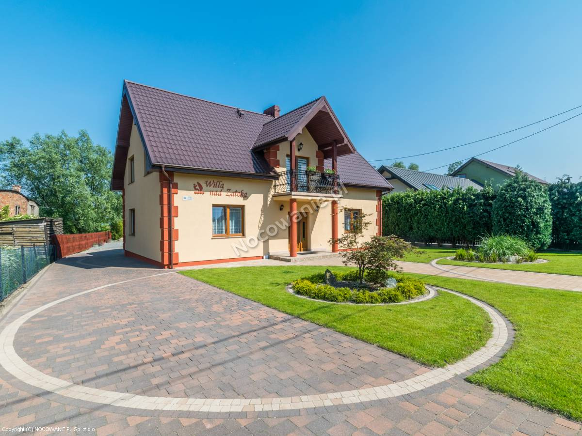 Dwupiętrowa willa z brązowym dachem, beżową elewacją i balkonem, otoczona zadbanym trawnikiem i brukowanym podjazdem z dekoracyjnym okręgiem.