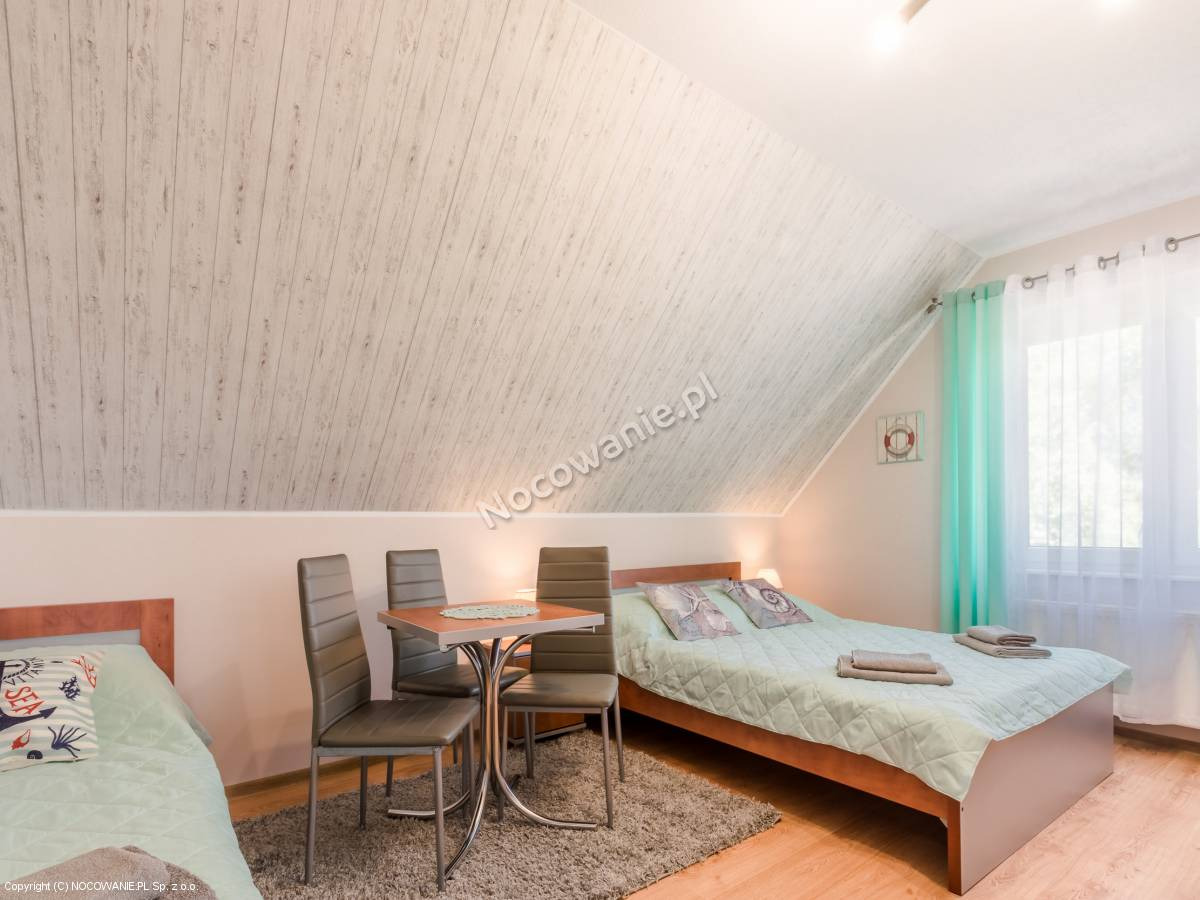 Pokój w pensjonacie na poddaszu z dwoma łóżkami, stołem i krzesłami, morskie akcenty dekoracyjne.