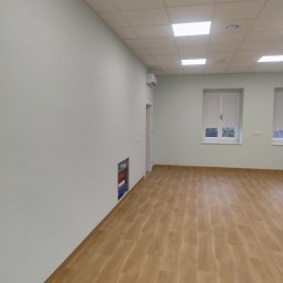 CatHouse - Jasne panele podłogowe w nowym, pomalowanym na jasno pomieszczeniu biurowym z oknami i sufitem kasetonowym. Widoczna instalacja elektryczna w ścianie.