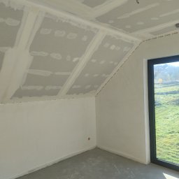 CatHouse - Poddasze z oknem po montażu płyt karton-gips, widoczne łączenia płyt i szpachlowanie. Świeża wylewka na podłodze. Widok na zewnątrz przez okno.